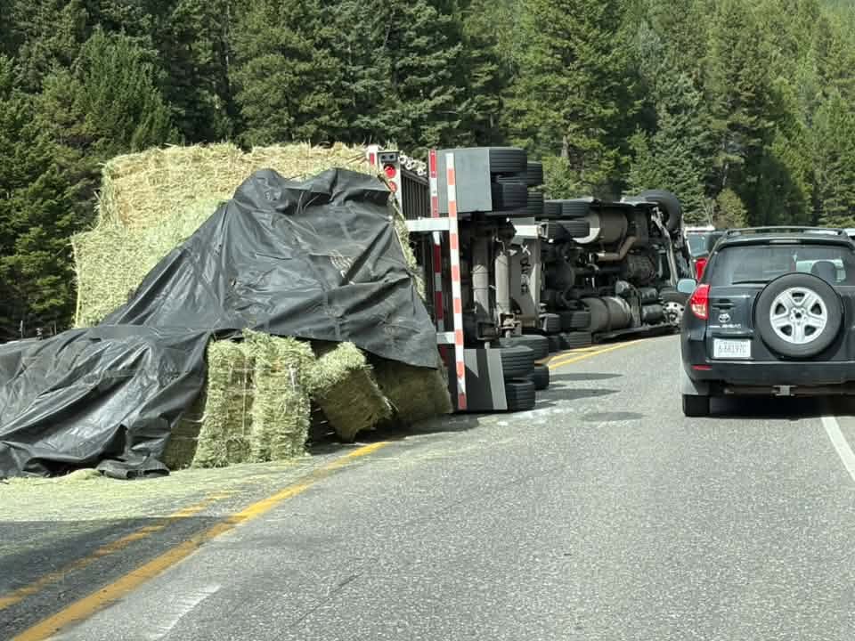 The flatbed on its side across the northbound lane of US-191, bales of hay spilled and half covered by a black tarp, with cars squeezing past in the other lane